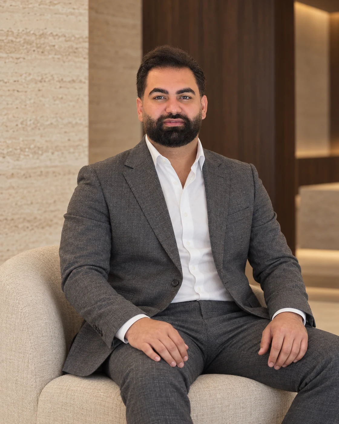 AI-assisted editorial portrait of Amin Alikhani seated in charcoal tailoring, with warm travertine and walnut surroundings